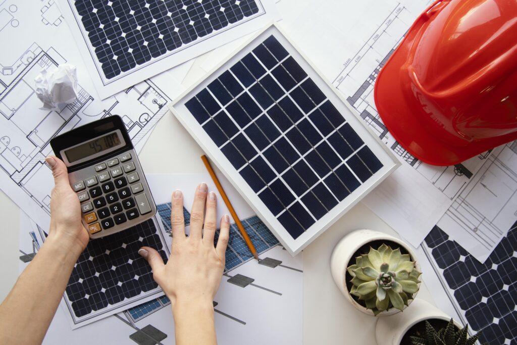 Mão apoiada na mesa junto com vários papéis e representação de um painel solar, com calculadora do lado, realizando um orçamento solar para projetos fotovoltaicos
